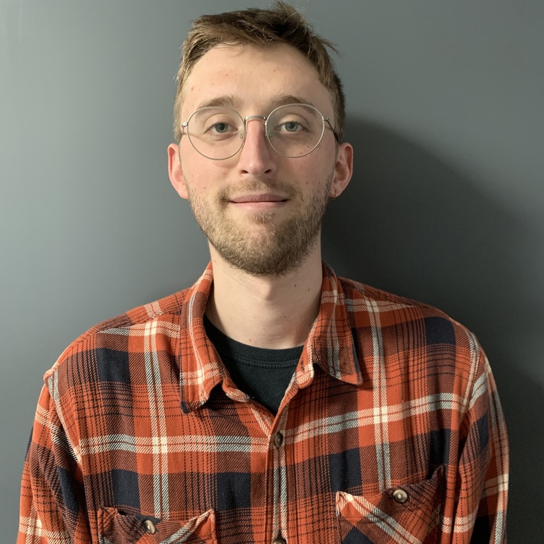 young man wearing red and yellow plaid button down shirt, short hair and glasses in front of gray wall 