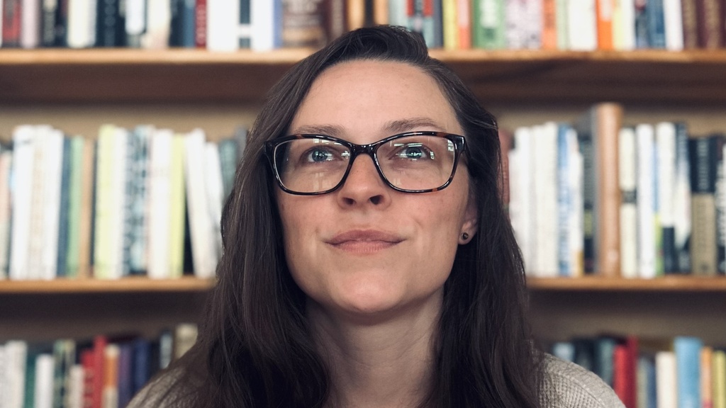 woman with long brown hair and glasses sitting in front of a shelf of books